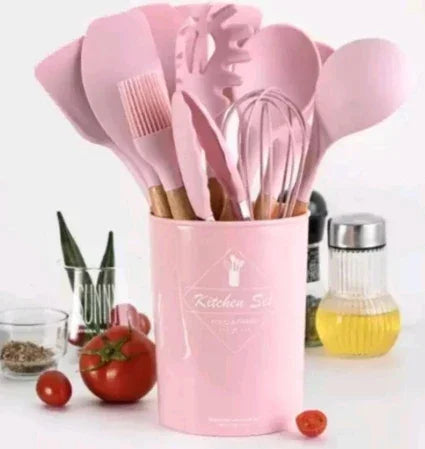 Conjunto de utensílios de cozinha rosa em pote, com tomates e temperos ao fundo.